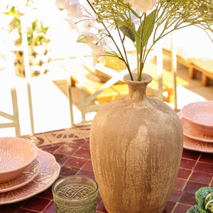 Peut inclure: Un vase en terre cuite à la finition rustique contient un bouquet de fleurs blanches sur une table en carreaux rouges avec des assiettes roses et vertes.