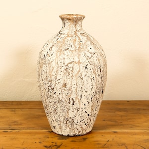 Peut inclure: Vase décoratif blanc cassé avec une finition vieillie. Le vase a un corps arrondi et un col étroit, avec un bord marron. La surface présente un aspect texturé avec des accents marron et noirs. Le vase est posé sur une surface en bois.