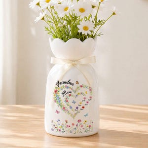 May include: White ceramic vase with a scalloped top, tied with a cream ribbon. The vase features a floral heart design with the words "Grandma Mom" and several names. The vase is filled with white daisy flowers.