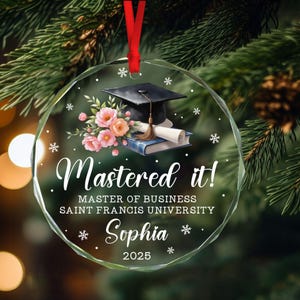 Peut inclure: Ornement de graduation en verre transparent avec un ruban rouge. L'ornement présente une toque noire, un diplôme et un bouquet de fleurs roses et pêche. Le texte indique "Mastered it!" et "Sophia 2025".