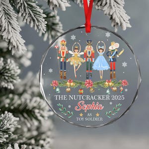 May include: A clear, faceted Christmas ornament with a red ribbon. The ornament features a Nutcracker ballet design with characters and the text "THE NUTCRACKER 2025" and "Sophia as TOY SOLDIER".