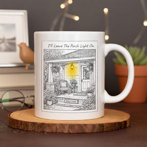 May include: White ceramic mug featuring a black and white illustration of a porch with a glowing light and the text "I'll Leave The Porch Light On." The mug is on a wooden coaster, with a blurred background including a small bird figurine and a potted plant.