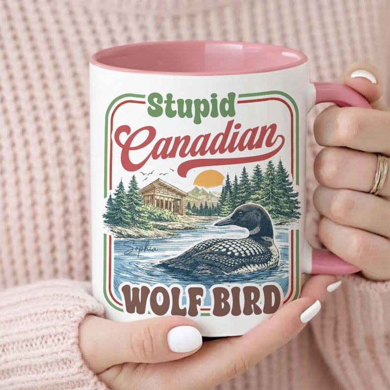 May include: White ceramic mug with a pink interior and handle. The mug features the text "Stupid Canadian WOLF-BIRD" with a graphic of a loon on a lake, trees, and a building. The mug is held by a person.