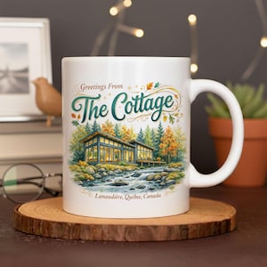 May include: White ceramic mug with the text "Greetings From The Cottage" and a scenic illustration of a modern cabin by a river. The mug has a curved handle and is on a wooden coaster.