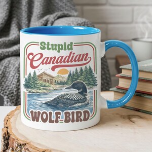 May include: White ceramic mug with a blue interior and handle. The mug features the text "Stupid Canadian WOLF-BIRD" with a graphic of a loon on a lake, trees, and a sunset. The mug is sitting on a wooden surface.
