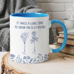Pode incluir: Caneca de cerâmica branca com interior e alça azuis. A caneca apresenta o texto "IT TAKES A LONG TIME TO GROW AN OLD FRIEND" com ilustrações florais e os nomes "Jessica" e "Sophia" em azul.