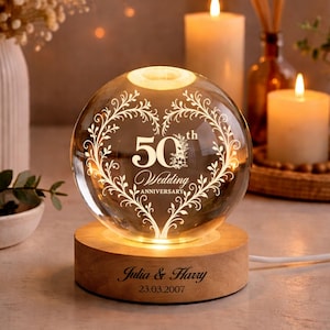 Könnte beinhalten: Eine klare Glaskugel mit leuchtendem Inneren, graviert mit "50. Hochzeitstag" und einem dekorativen Herzdesign. Die Kugel steht auf einem Holzsockel mit eingravierten Namen und einem Datum. Kerzen und Dekoration im Hintergrund.