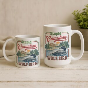 May include: Two white ceramic mugs with the text "Stupid Canadian WOLF BIRD" in a retro design. The design includes a loon on a lake, trees, and a sunset. One mug is smaller than the other.