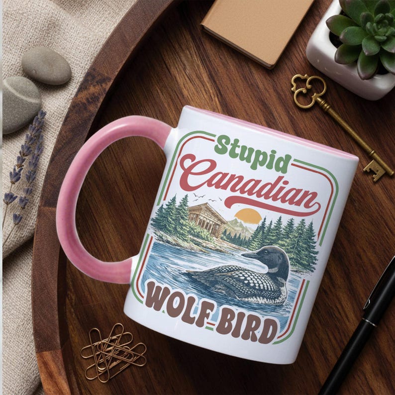 May include: White ceramic mug with a pink handle and rim. The mug features the text "Stupid Canadian Wolf-Bird" with a graphic of a loon on a lake, trees, and a building. The design is in red, green, and brown.