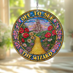 Puede incluir: Adorno redondo de vidriera con el texto "OFF TO SEE THE WIZARD". El diseño presenta un camino de baldosas amarillas que conduce a un castillo, rodeado de flores y follaje. Una cadena plateada está adjunta para colgar.