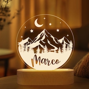 May include: A personalized night light featuring a mountain and forest design with a crescent moon and stars. The name "Marco" is displayed in a script font. The light sits on a wooden base.