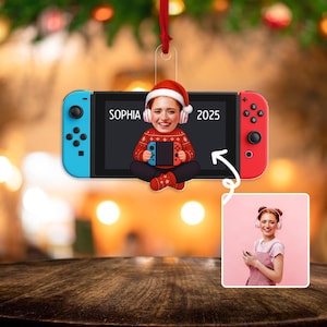 May include: A Christmas tree ornament in the shape of a games console. The ornament has a picture of a person wearing a Santa hat and holding a game controller. The name "SOPHIA" and the year "2025" are printed on the screen. The ornament is blue and red.