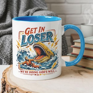 Puede incluir: Taza de cerámica blanca con interior y asa azules. La taza presenta un gráfico humorístico con el texto "GET IN LOSER" sobre una ilustración de una ballena y una persona en el agua. La frase "WE'RE DOING GOD'S WILL" está debajo.