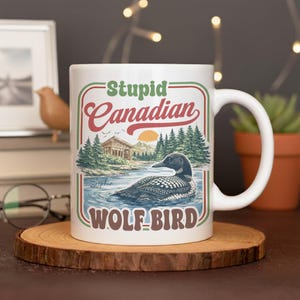 Puede incluir: Taza de cerámica blanca con asa, con el texto "Stupid Canadian WOLF-BIRD" en un diseño retro. La taza presenta una ilustración detallada de un somorgujo en un lago con un fondo escénico de árboles y un atardecer.