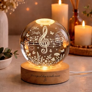 Puede incluir: Una esfera de cristal iluminada con notas musicales, mariposas y diseños florales, sobre una base de madera. La esfera está grabada con el nombre "Jenifer" y la frase "It's a good day to make music."