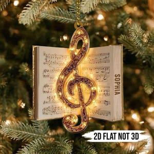 以下が含まれることがあります： 小さなライトが付いた金色のト音記号のクリスマスオーナメントが、開いた楽譜の本の前に吊り下げられています。本のページが見え、右ページには「SOPHIA」という名前が印刷されています。「2D FLAT NOT 3D」というテキストが下にあります。