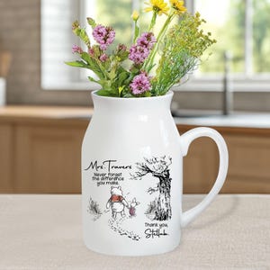 May include: White ceramic vase with a Winnie the Pooh and Piglet design. The vase has a handle and features the text "Mrs. Travers Never forget the difference you make. Thank you, Stella."
