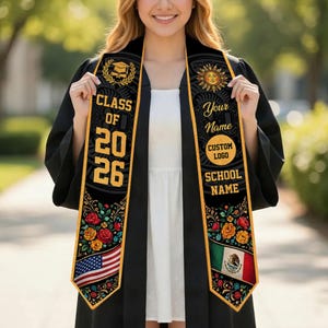 Op de afbeelding: Zwarte afstudeer sjerp met gouden rand en tekst. De sjerp heeft de tekst "CLASS OF 2026", "Your Name", "Custom Logo" en "School Name". Bloemen- en vlagontwerpen zijn ook aanwezig. De sjerp wordt gedragen over een witte jurk en een zwarte toga.