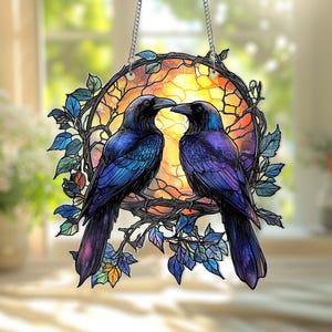 Puede incluir: Colgante decorativo de vidriera con dos cuervos negros posados en una rama. Las aves tienen plumas iridiscentes azules y moradas, sobre un fondo cálido color puesta de sol. El diseño está enmarcado por un borde circular con hojas y vides.