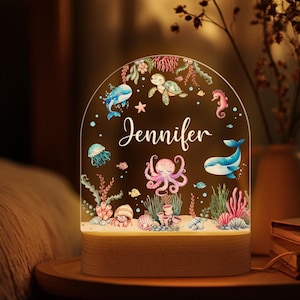 Puede incluir: Una luz nocturna personalizada con una base de madera y una cúpula de acrílico transparente. La cúpula presenta una escena submarina caprichosa con una ballena, un pulpo, una tortuga, una estrella de mar y un caballito de mar. El nombre "Jennifer" está escrito en blanco en la cúpula.