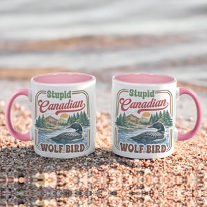 May include: Two white ceramic mugs with pink handles and interiors. Each mug features a vintage-style graphic with the text "Stupid Canadian" above an illustration of a loon on a lake, with the words "WOLF BIRD" below.
