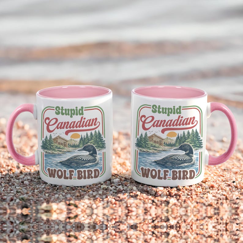 May include: Two white ceramic mugs with pink handles and interiors. Each mug features a vintage-style graphic with the text "Stupid Canadian" above an illustration of a loon on a lake, with the words "WOLF BIRD" below.