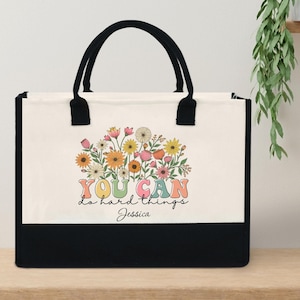 Peut inclure: Un sac fourre-tout en toile avec une base et des poignées noires. La partie supérieure est blanche, affichant un motif floral et le texte "YOU CAN do hard things Jessica" en lettres colorées. Le sac est conçu pour transporter des articles.