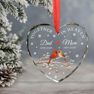 以下が含まれることがあります： 「TOGETHER IN HEAVEN」と「Dad」と「Mom」の文字が入ったハート型のガラスオーナメント。赤い鳥が枝に止まり、雪の結晶と日付が描かれています。吊り下げ用の赤いリボンが付いています。