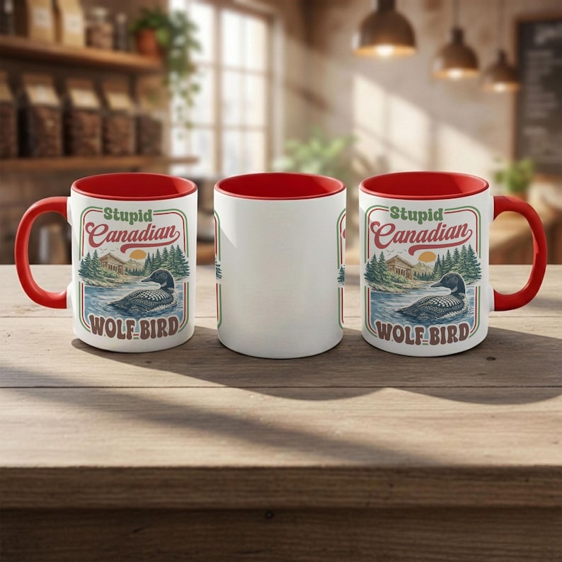 May include: Three white ceramic mugs with red interiors and handles are displayed on a wooden surface. Each mug features the text "Stupid Canadian" above an illustration of a loon on a lake. The mugs are a humorous gift idea.