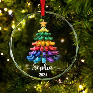 Puede incluir: Un adorno de Navidad de vidrio con un árbol de Navidad de colores arcoíris hecho de zuecos coloridos. El adorno está personalizado con el nombre "Sophia" y el año "2024".