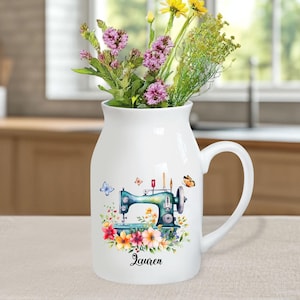 May include: White ceramic pitcher with a handle, featuring a vintage sewing machine design with colorful flowers and butterflies. The pitcher has the name "Lauren" printed on the bottom.