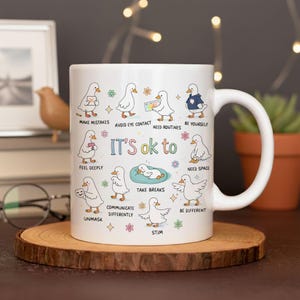 May include: White ceramic mug featuring cartoon ducks with various affirmations like "It's ok to make mistakes" and "Be yourself." The mug has a curved handle and is set on a wooden coaster.