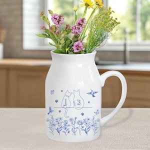 May include: White ceramic vase with a handle, decorated with blue cat silhouettes, flowers, butterflies, and hummingbirds. The vase holds a bouquet of colorful flowers. The names "Karen" and "Be" are written on the cats. A charming and decorative piece.