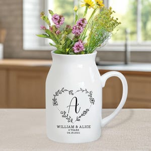 以下が含まれることがあります： 白いセラミックの花瓶、取っ手付き、カラフルな花でいっぱい。花瓶には、黒いハート型の花の模様があり、中に文字「A」があります。ハートの下には「WILLIAM & ALICE 4 YEARS 04.18.2021」と書かれています。