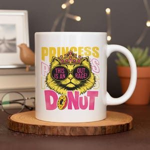 以下が含まれることがあります： 白いセラミックマグカップ。サングラスと王冠をつけた漫画の猫が描かれています。「PRINCESS」と「DONUT」の文字がピンクと黄色で書かれており、猫のサングラスには「THIS IS AN」と「OUT RAGE！」のフレーズが書かれています。