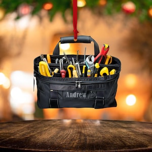 Peut inclure: Ornement de Noël en forme de sac à outils noir rempli d'outils divers, dont des clés, des tournevis et des pinces. Le nom "Andrew" est imprimé sur le devant. L'ornement est suspendu à un ruban rouge, avec un arrière-plan flou.