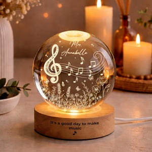 Puede incluir: Una esfera de vidrio transparente con notas musicales, flores y mariposas grabadas, iluminada desde el interior. Se apoya sobre una base de madera con la frase "It's a good day to make music." El nombre "Mrs. Annabelle" también está grabado.