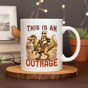 以下が含まれることがあります： サングラスとマントを着た猫が恐竜に乗っているイラストが描かれた白いセラミックマグカップ。「THIS IS AN OUTRAGE」の文字が茶色でプリントされています。マグカップには大きなハンドルが付いています。