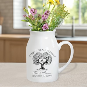 May include: White ceramic pitcher with a handle, filled with colorful flowers. The pitcher is decorated with a black tree design, a heart, and the words "Happy 9th Anniversary", "Ben & Caroline", and "Rooted in Love".