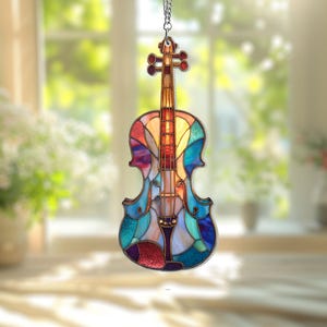 Könnte beinhalten: Ein farbenfrohes Geigenornament aus Glas hängt an einer silbernen Kette. Die Geige zeigt ein Mosaik aus Blau-, Grün-, Rot- und Gelbtönen. Der Korpus des Instruments ist dunkelbraun umrandet, mit einem verschwommenen Fenster im Hintergrund.