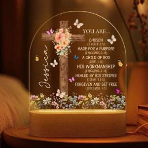 May include: A wooden cross with a floral design and butterflies is inside a clear glass dome with a light base. The text "YOU ARE... CHOSEN (1 PETER 2:9) MADE FOR A PURPOSE (EPHESIANS 2:10) A CHILD OF GOD (JOHN 1:12) HIS WORKMANSHIP (EPHESIANS 2 10) HEALED BY HIS STRIPES (ISAIAH 53:5) FORGIVEN AND SET FREE (EPHESIANS 1:7)" is printed on the glass. The name "Jessica" is written on the side of the dome.