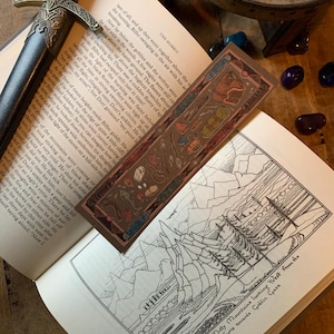 Puede incluir: Primer plano de un marcapáginas con una ilustración detallada de una mochila y otros equipos de campamento. El marcapáginas está hecho de material marrón texturizado con un borde dorado. El texto "Remarkable" está impreso en el marcapáginas. El marcapáginas está colocado en un libro con una ilustración en blanco y negro de una cordillera y el texto "The Misty Mountains looking West from the Eyrie towards Goblin Gate".