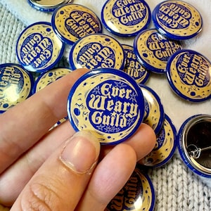 Può includere: Una collezione di bottoni rotondi blu e oro con la scritta "Ever Weary Guild". I bottoni presentano un design a mezzaluna e stelle. Alcuni bottoni sono rivolti verso l'alto, mentre altri mostrano i retro con le spillette. I bottoni sono sparsi su una superficie chiara e strutturata.