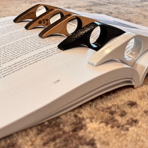 May include: Four different book page holders, two are made of wood and two are made of plastic. The wooden holders are brown and have a natural wood grain. The plastic holders are black and white. The holders are all in the shape of a curved arch.