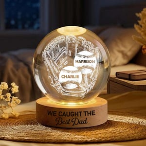 May include: A clear glass sphere with a baseball glove and two baseballs etched inside. The baseballs are labeled "Harrison" and "Charlie." The sphere sits on a wooden base with the text "WE CAUGHT THE Best Dad."
