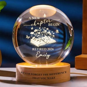 May include: A clear glass sphere lamp on a cylindrical wooden base. The sphere features an etched design of an open book with flowers and butterflies, with the text A NEW CHAPTER BEGIN, RETIRED 2026, and Emily. The base reads NEVER FORGET THE DIFFERENCE THAT YOU MAKE.
