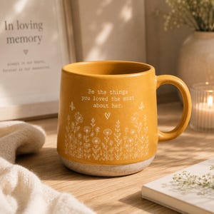 May include: A mustard yellow ceramic mug featuring a white line art illustration of wildflowers and butterflies. The text "Be the things you loved the most about her." is printed in white above a small heart icon. The base has a speckled texture.