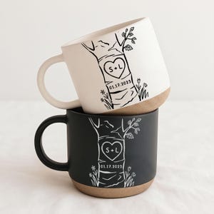 May include: Two ceramic mugs stacked vertically, one white and one black, each featuring a black or white line drawing of a tree trunk with a heart containing the letters S+L and the date 01.17.2023, with a natural tan base on both mugs.