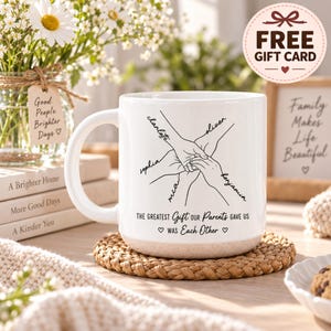 Peut inclure: Un mug en céramique blanche avec une base mouchetée présentant un dessin au trait noir de quatre mains jointes. Les prénoms Charlotte, Oliver, Sophia, Mia et Benjamin sont inscrits près des mains. Le texte indique : THE GREATEST GIFT OUR PARENTS GAVE US WAS EACH OTHER.