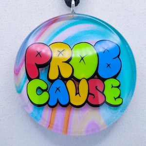Może przedstawiać: Naszyjnik z okrągłą zawieszką z napisem "PROB CAUSE" w kolorowych literach bąbelkowych. Zawieszka ma wirujące tło w stylu tie-dye w odcieniach niebieskiego, różowego i żółtego. Naszyjnik ma czarny sznurek.